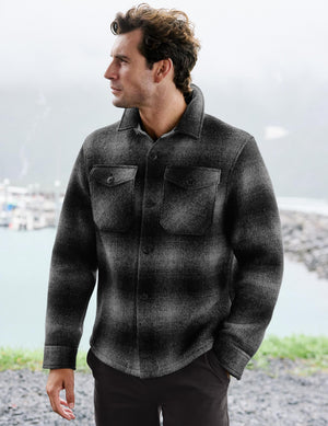 A man wearing a dark gray and black plaid button-up shirt jacket, standing outdoors with a blurred background of water and rocks.