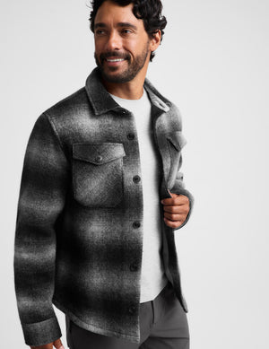 A man wearing a dark gray and black textured plaid overshirt over a white t-shirt, photographed against a solid light gray background.