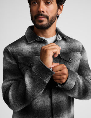 A man wearing a charcoal gray textured overshirt jacket over a light gray t-shirt, adjusting the collar and cuff of the jacket against a plain light gray background.