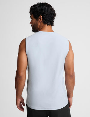 A man viewed from the back wearing a light blue sleeveless muscle tank top and black pants, set against a plain light gray studio background.