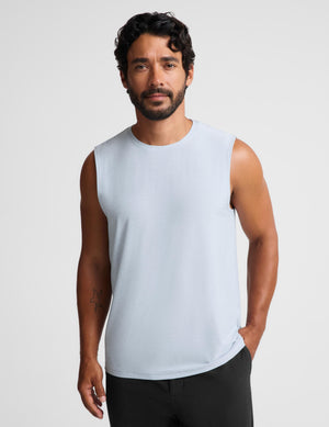 A man wearing a light blue sleeveless crewneck tank top and black pants, standing against a light gray studio background.