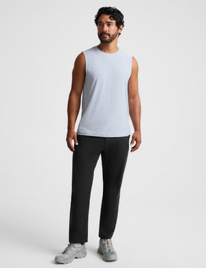 A man modeling a light blue sleeveless muscle tank top paired with black pants and gray sneakers, standing against a plain light gray studio background.