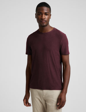 A man wearing a short-sleeve crewneck t-shirt in maroon and beige trousers, standing against a light gray studio background.