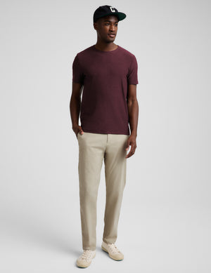 A man wearing a short-sleeve crewneck t-shirt in maroon and beige trousers, standing against a light gray studio background.