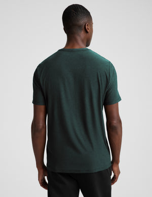A man wearing a short sleeve green t-shirt and black pants against a plain studio background. 