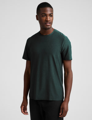 A man wearing a short sleeve green t-shirt and black pants against a plain studio background. 