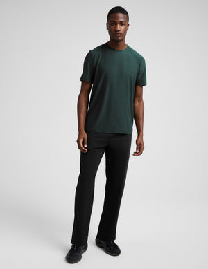 A man wearing a short sleeve green t-shirt and black pants against a plain studio background. 