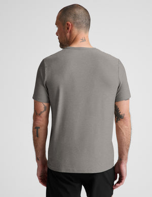 A man viewed from the back wearing a plain heather gray short-sleeve t-shirt and black pants, standing against a light gray studio background.