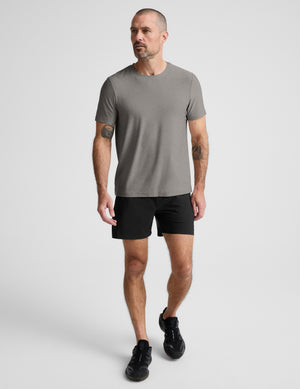 A man wearing a plain gray short-sleeve crewneck t-shirt, black athletic shorts, and black sneakers, standing against a light gray studio background.