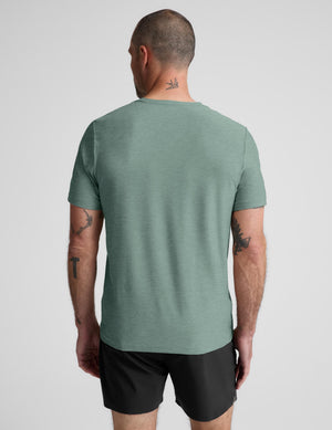 A man viewed from behind wearing a heathered sage green short-sleeve t-shirt and black athletic shorts against a light gray studio background.