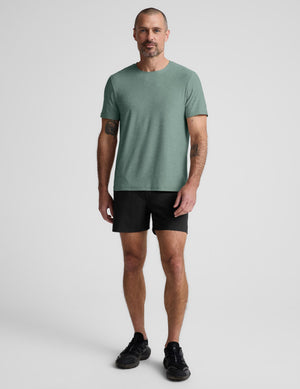 A man modeling a sage green crew-neck t-shirt and black athletic shorts against a light gray studio background.