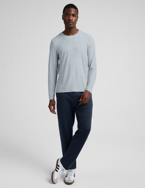 A man wearing a light gray long-sleeve crewneck shirt and dark navy pants, standing against a light gray studio background.