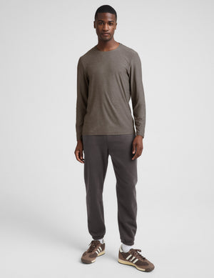 A man wearing a brown long sleeve crew neck shirt and dark gray pants against a plain studio background. 