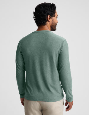 A man viewed from the back wearing a heathered sage green long-sleeve crewneck shirt and beige pants, set against a plain light gray studio background.