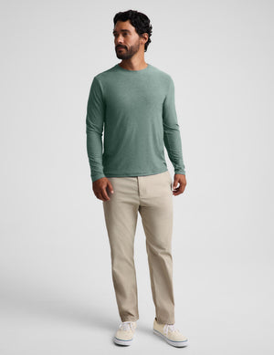 A man wearing a sage green long-sleeve crewneck shirt, beige trousers, and off-white sneakers, standing against a light gray studio background.