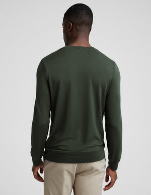 A man wearing a dark olive green long-sleeve crewneck sweater, paired with beige trousers, photographed against a neutral light gray background.