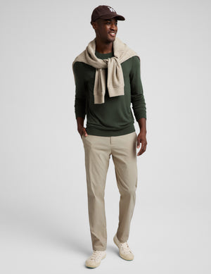 A man modeling a dark green crewneck sweater, beige trousers, a beige knit scarf, and a dark brown baseball cap against a light gray studio background.