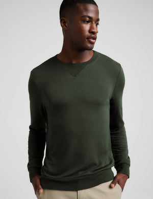 A man wearing a dark olive green long-sleeve crewneck sweater, paired with beige trousers, photographed against a neutral light gray background.