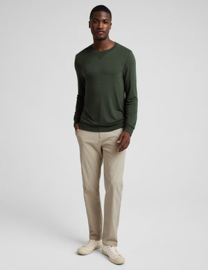 A man wearing a dark olive green long-sleeve crewneck sweater, paired with beige trousers, photographed against a neutral light gray background.