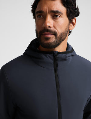 A close-up shot of a man wearing a dark navy blue zip-up hooded jacket against a light gray background.
