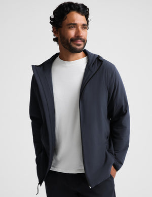 A man wearing a dark navy lightweight zip-up hooded jacket over a white crewneck t-shirt, standing against a light gray studio background.