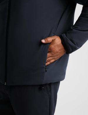 Close-up of a person wearing a dark navy jacket, with one hand inserted into a zippered pocket, against a light gray background.