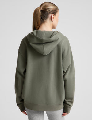 A woman viewed from the back wearing an oversized olive green pullover hoodie, paired with light gray leggings, against a solid light gray studio background. | womens