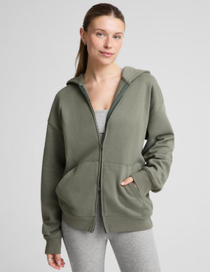 A woman wearing an oversized olive green zip-up hoodie with a front pocket, paired with gray leggings, against a light gray studio background. | womens