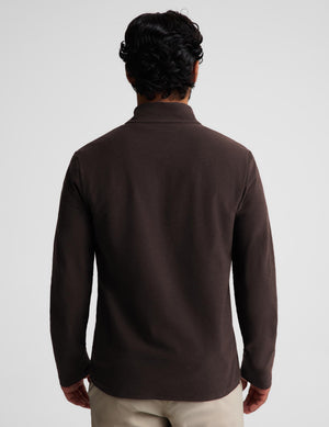 A man wearing a dark brown quarter-zip long-sleeve knit pullover over a white t-shirt and khaki pants, standing against a light gray studio background.