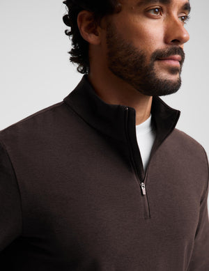 A man wearing a dark brown quarter-zip long-sleeve knit pullover over a white t-shirt and khaki pants, standing against a light gray studio background.