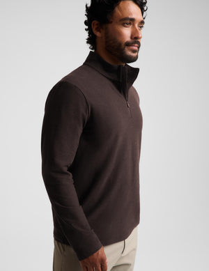 A man wearing a dark brown quarter-zip long-sleeve knit pullover over a white t-shirt and khaki pants, standing against a light gray studio background.