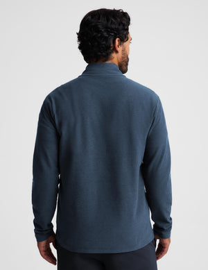 A man viewed from the back wearing a dark slate blue long-sleeve quarter-zip fleece, set against a plain light gray studio background.