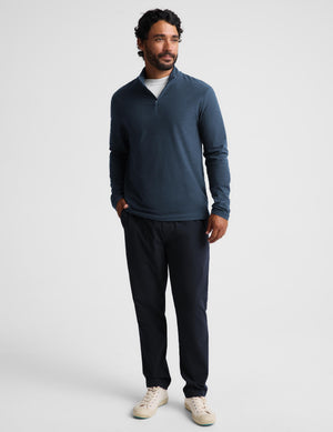 A man modeling a dark slate blue quarter-zip long-sleeve shirt and dark navy pants against a light gray studio background.