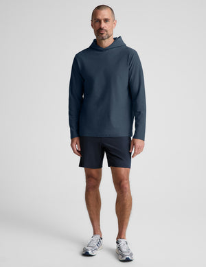 A man wearing a dark slate blue long-sleeve athletic hoodie and matching dark shorts, standing against a light gray studio background.