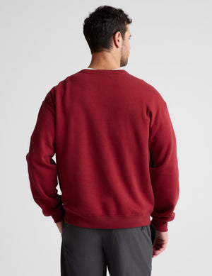 A man viewed from behind wearing a solid dark red crewneck sweatshirt and dark gray pants, set against a plain light gray studio background.