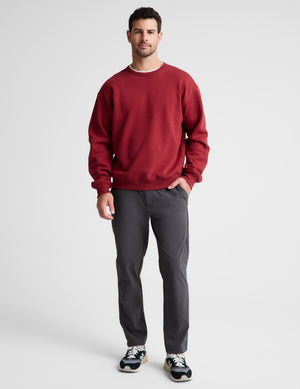 A man wearing a red crewneck sweatshirt, dark gray pants, and sneakers, standing against a light gray studio background.