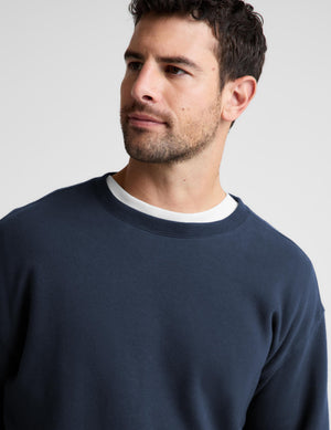 A close-up of a man wearing a navy blue crewneck sweatshirt layered over a white t-shirt, photographed against a light gray studio background.
