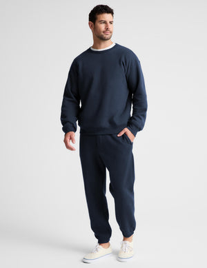 A man wearing a navy blue crewneck sweatshirt with a contrasting white inner collar, paired with matching navy pants, photographed against a light gray studio background.