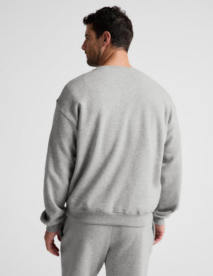 A man wearing a light gray heather crewneck sweatshirt and matching sweatpants, shown from a rear three-quarter view against a plain light gray studio background.