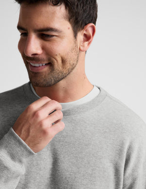 A close-up of a man wearing a light gray crewneck sweatshirt over a white undershirt, set against a plain light gray background.