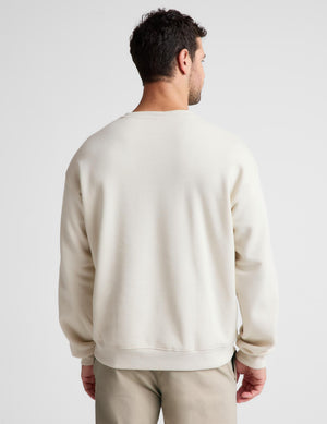 A man viewed from behind wearing a cream-colored crewneck sweatshirt and khaki pants against a light gray studio background.