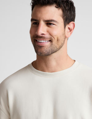 A close-up shot of a smiling man wearing a light cream crewneck sweatshirt against a plain light gray background.