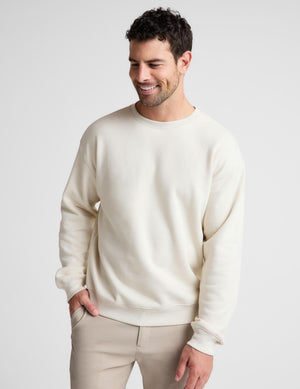 A man wearing a cream-colored crewneck sweatshirt and beige trousers, standing against a light gray studio background.