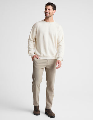 A man wearing a cream crewneck sweatshirt, beige trousers, and dark brown boots, standing against a light gray studio background.