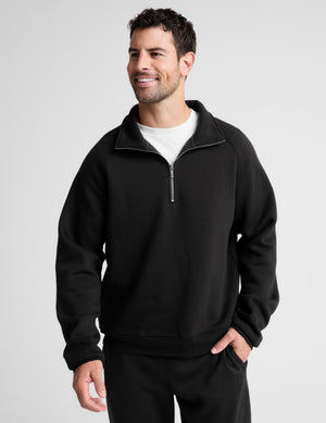 A man wearing a black quarter-zip sweatshirt with a mock neck over a white t-shirt, standing against a light gray studio background.