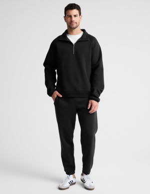 A man modeling a black half-zip sweatshirt and matching black sweatpants, paired with white sneakers, against a light gray studio background.