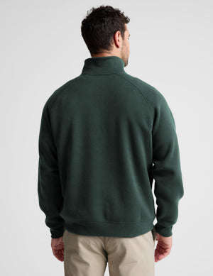 A man viewed from the back wearing a dark green quarter-zip sweatshirt and beige pants, set against a plain light gray studio background.