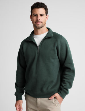 A man wearing a dark green quarter-zip sweatshirt over a white t-shirt and khaki pants, standing against a light gray studio background.
