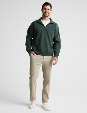 A man modeling a dark green quarter-zip fleece pullover, paired with beige trousers and white sneakers, against a light gray studio background.