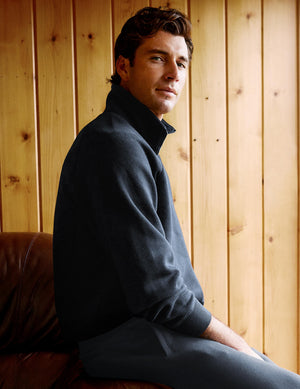 A man wearing a dark charcoal gray mock-neck long-sleeve sweater and matching gray trousers, seated on a brown leather couch in front of a light wood-paneled wall.
